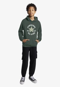 Žalias džemperis su balta "Converse All Star" logotipu ir žvaigždės detalė, derinamas su juodomis cargo kelnėmis ir juodai baltais sportbačiais.