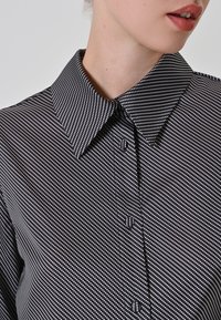 Camicia a pois bianco e nero con colletto a punta, chiusura a bottoni e tessuto testurizzato. Il motivo è diagonale con piccoli pois distinti.