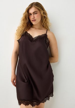 Vrouw met krullend blond haar die een mouwloze zwarte satijnen slipjurk met kanten afwerking draagt, staand tegen een effen witte achtergrond.