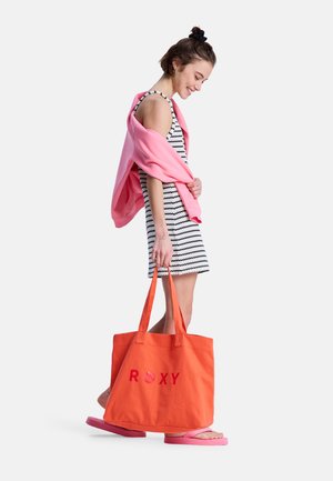 Young woman in striped sleeveless dress and pink flip-flops carries large orange tote bag and pink sweater draped over shoulders.