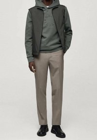 Homme portant un sweat à capuche vert, un gilet sans manches foncé, un pantalon beige et des chaussures à lacets noires, debout devant un fond uni.