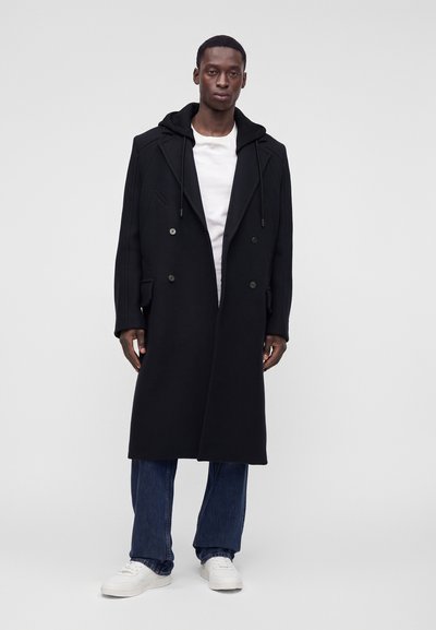 Homme portant un long manteau noir croisé avec capuche, t-shirt blanc, jean bleu et baskets blanches, debout devant un fond uni.