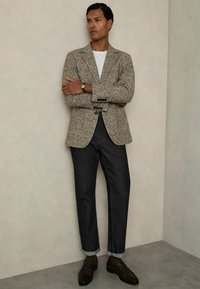 Blazer tejido marrón y beige con textura y dos botones, camiseta blanca, pantalones de mezclilla oscuros con dobladillos, y mocasines marrones, todo ello sobre un fondo neutro.