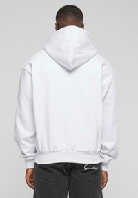 Sudadera blanca con capucha, de corte relajado, con puños y dobladillo acanalados. Textura suave, con un bolsillo tipo canguro y detalle del logo en la parte trasera.