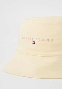 Gul bøttehatt laget av stoff, med brodert "TOMMYJEANS" tekst i rosa og en liten tri-farge logo aksent. Teksturert kant.