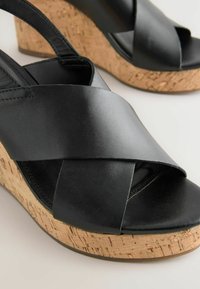 I sandali con zeppa in pelle nera presentano fasce incrociate con un design in sughero, offrendo un contrasto di texture e una silhouette elegante e moderna.