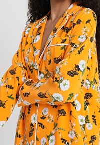 Orangefarbene Schlafanzugbluse mit schwarz-weißen Blumenmustern, ausgestattet mit einem Kragen, langen Ärmeln und weißen Paspeln an den Rändern.
