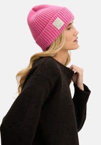 Różowa dziergana czapka z prążkowaną teksturą i podwiniętym mankietem, z małą metką marki. Noszona z brązowym swetrem.
