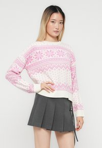Woman wearing a white and pink patterned sweater and a gray pleated skirt with a side tie, posing with one hand on her hip.