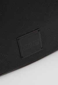 Černá texturovaná taška s embosovaným vzorem "Versace Jeans Couture" a obdélníkovou černou etiketou s výrazně zobrazeným názvem značky.