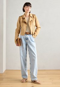 Tan leather motorcycle jacket with silver zippers, beige top, light blue high-waisted jeans, and brown sandals against a neutral background.