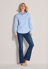 Hellblaues Hemd mit weißen Streifen, lange Ärmel und abgerundetem Saum, kombiniert mit dunkelblauen Schlaghosen und beige gefütterten Schuhen.