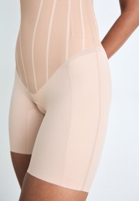Body beige avec un design ajusté, comportant des lignes et des coutures verticales. Texture lisse et longueur mi-cuisse.