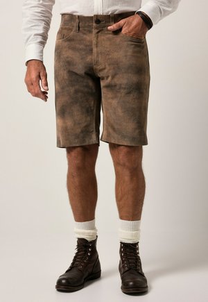 Man draagt bruine suède shorts, witte sokken, donkerbruine veterlaarzen en een wit overhemd met lange mouwen, met één hand in de zak.