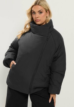 Mujer con chaqueta acolchada negra con cremallera asimétrica y cuello alto, de pie con una mano en el bolsillo frente a un fondo claro.