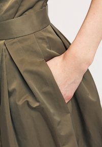 Oliivinvihreä mekko, jossa on istuva vyötärö ja laskostettu materiaali. Lähikuva kädestä, joka on asetettu sivutaskuun, korostaen tekstuuria ja kangasta.