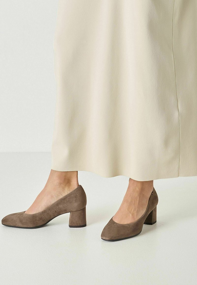 Jambes portant une jupe midi beige et des chaussures en daim marron à talons blocs avec des bouts arrondis sur un sol blanc lisse.