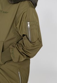 Olijfgroene bomberjack met een bontgevoerd capuchon, ritssluiting op de schouder, elastische manchetten en geborduurde vleugeldetails op de mouw.