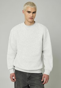 Sweatshirt gris clair avec un col rond, des poignets côtelés et un ourlet côtelé. Le tissu a une apparence texturée et douce, complété par un pantalon gris foncé.