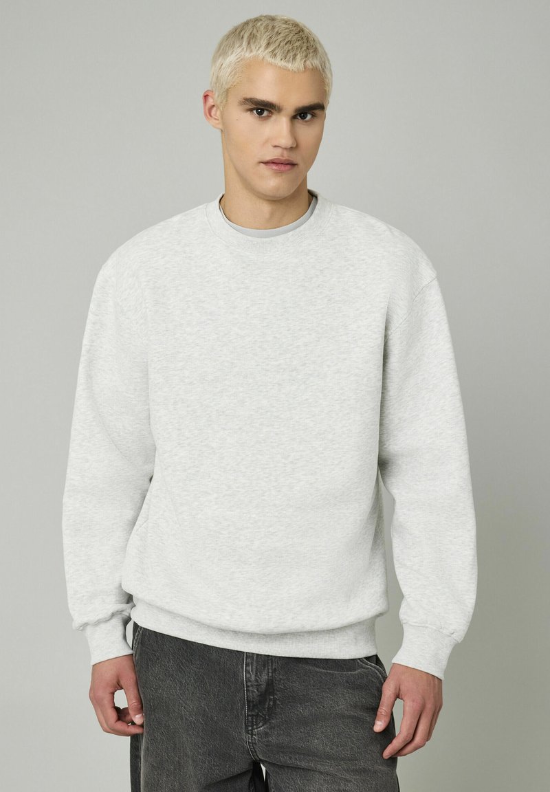 Sweatshirt gris clair avec un col rond, des poignets côtelés et un ourlet côtelé. Le tissu a une apparence texturée et douce, complété par un pantalon gris foncé.