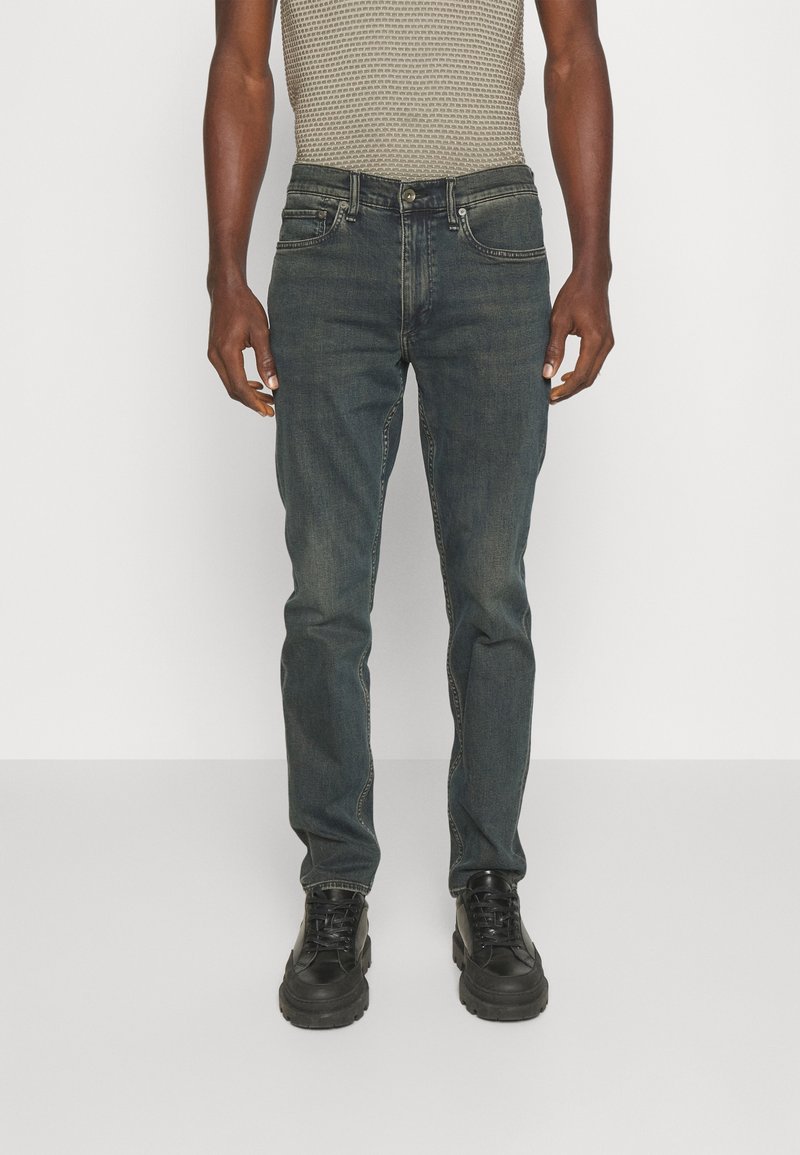 Jeans de mezclilla oscura con corte recto, que presentan costuras en contraste y un diseño de cinco bolsillos, combinados con una blusa texturada gris y botas negras.