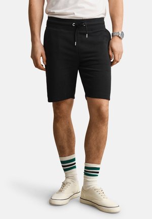 Homme portant un short noir à cordon, des chaussettes blanches avec des rayures vertes et noires, des baskets blanches et une chemise blanche, debout sur un fond uni.
