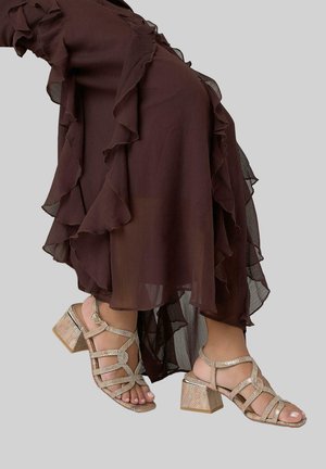 Pies con sandalias beige de tacón bloque con tiras, combinadas con una falda marrón oscuro transparente con volantes.