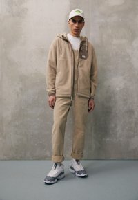 Beige fleecetröja med en brun lapp, beige overaller och gråvit sneakers. Modellen bär en ljus keps med en grön logotyp.