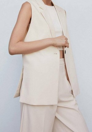 Femme portant un blazer beige sans manches, un pantalon large assorti et un top court blanc, ajustant le blazer d'une main.