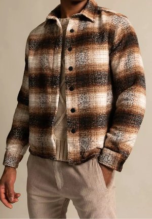 Homme portant une veste boutonnée à carreaux marron, noire et blanche sur un pull en maille beige et un pantalon en velours côtelé beige clair, main dans la poche.
