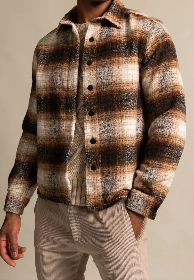 Homme portant une veste boutonnée à carreaux marron, noire et blanche sur un pull en maille beige et un pantalon en velours côtelé beige clair, main dans la poche.