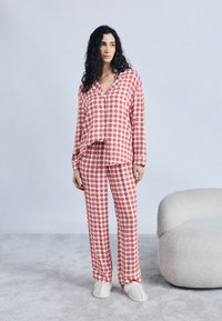 Γυναίκα που στέκεται με κόκκινη και άσπρη πιτζάμα καρό με σχέδια καρδιών, φορώντας άσπρες παντόφλες, κοντά σε μια κρεμ πολυθρόνα πάνω σε γκρι χαλί.