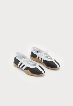 Chaussures basses noires et blanches avec une semelle en caoutchouc, présentant trois rayures horizontales, un bout arrondi et un système de lacets.