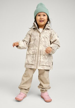 Junges Kind trägt eine mintgrüne Strickmütze, eine beige gemusterte Jacke, beige Hosen und rosa Slipper, steht vor einem einfarbigen hellen Hintergrund.