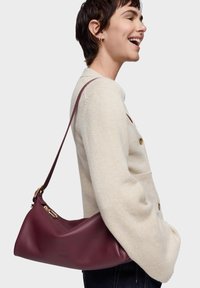 Bolso de hombro de cuero burdeos con forma curva, una sola correa y detalles en hardware dorado. Textura suave con detalle de logotipo.