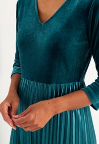Tiefgrünes Samtkleid mit V-Ausschnitt, drei Viertel langen Ärmeln und plissiertem Rock. Glatte Textur mit sanftem Glanz und definierten Falten an der Taille.