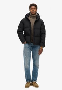 Giacca a vento nera con cappuccio, caratterizzata da un design trapuntato. Indossata sopra un dolcevita marrone, abbinata a jeans azzurri chiari e scarpe beige.