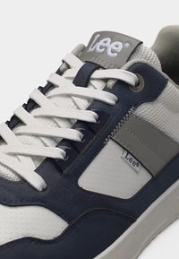 Λευκό, σκούρο μπλε και γκρι αθλητικό παπούτσι Lee με λευκά κορδόνια, ύφασμα από δίχτυ, και ετικέτες με το λογότυπο στη γλώσσα και στην πλαϊνή πλευρά.