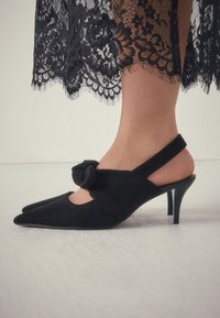 Escarpins slingback en daim noir avec un bout pointu et un détail noué sur l'empeigne. Ils présentent un talon haut et fin ainsi qu'un design ajouré.