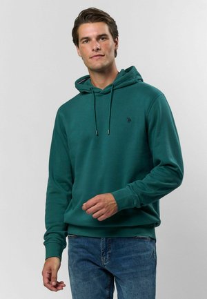 Sweat à capuche teal avec cordon de serrage, arborant un petit logo sur la poitrine. Texture douce et poignets côtelés, associé à un jean bleu.