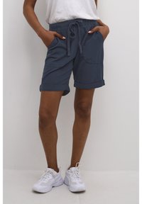 Pantalones cortos azul marino con cintura ajustable, puños enrollados y bolsillos laterales. La tela parece ligera y texturizada. Combinados con zapatillas blancas.