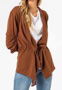 Brun omwrap cardigan i mjukt tyg. Har knytmidja, långa ärmar och en avslappnad passform. Matcher med en vit t-shirt och blå jeans.