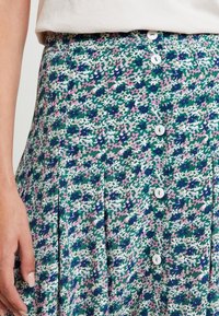 Blumengemusterter Rock in Grün mit rosa und weißen Akzenten. Verfügt über eine Knopfleiste, Falten und einen leichten, strukturierten Stoff.