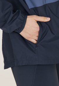 Giacca a vento navy con finitura testurizzata, dotata di un pannello blu superiore, polsini elastici e una tasca con zip sul lato.