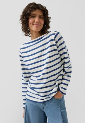 Frau mit lockigem Haar, die ein weiß-blau gestreiftes Langarmshirt und hellblaue Jeans trägt, lächelt mit einer Hand in der Tasche.