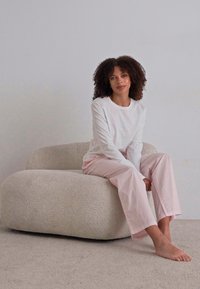 Lättviktig vit långärmad skjorta och mjuka rosa vida byxor. Modell som sitter på en rundad, krämfärgad texturerad stol. Neutral bakgrund.