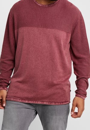 Mand iført en langærmet, farveopdelt rød og vinrød tekstureret sweatshirt og lysegrå jeans, stående mod en ensfarvet baggrund.