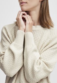 Pull en maille couleur crème avec des motifs en losanges texturés et des poignets côtelés. Les manches sont volumineuses et légèrement surdimensionnées.