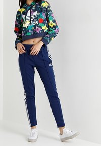 Felpa cropped di colore blu scuro con vivaci motivi floreali e logo Adidas bianco; abbinata a pantaloni tapered blu navy con strisce bianche.