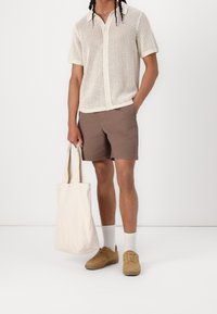 Chemise à manches courtes en maille crème avec un devant boutonné, associée à un short marron. Sac cabas beige et baskets marron clair avec des chaussettes blanches à la cheville.